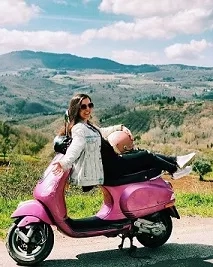 A woman is riding a pink Vespa on a road with mountains in the background.