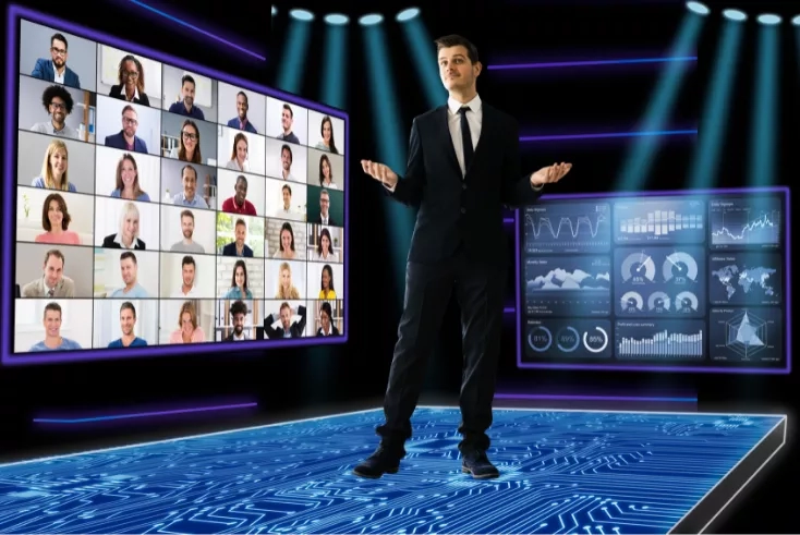 A man stands in front of a screen with people and a computer monitor.