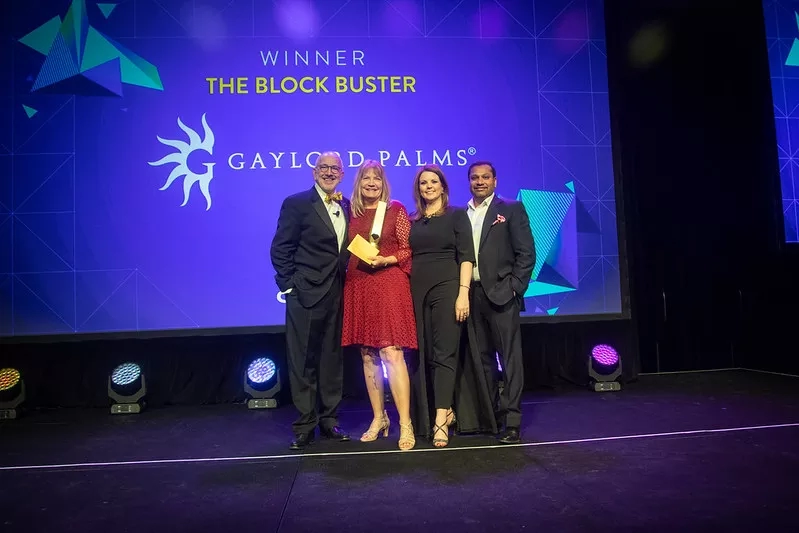 Four people stand on stage holding an award, with a screen behind them reading "Winner The Block Buster".