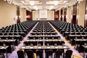 Large meeting room with black chairs and tables, a white screen, and a wooden podium.