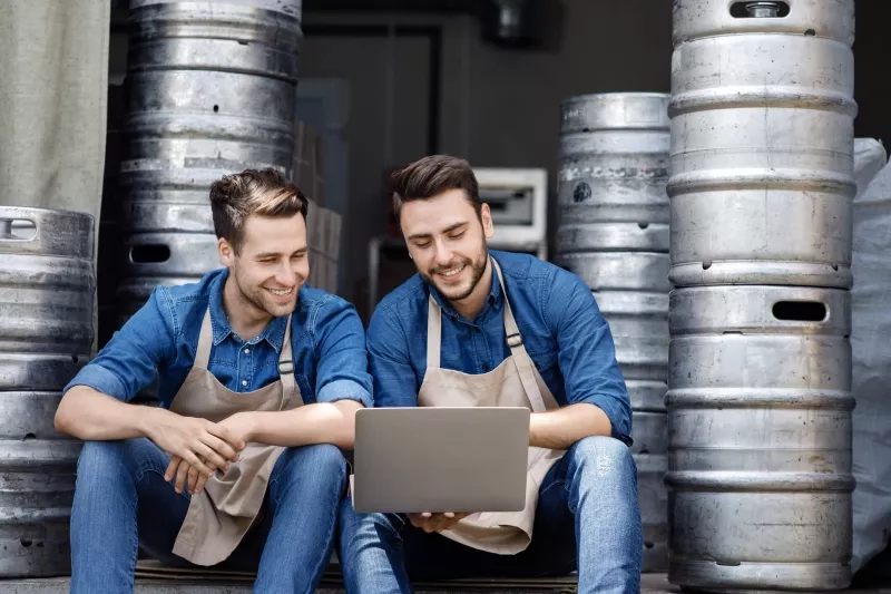 Two men in aprons using a laptop, sitting next to a wall of beer kegs.