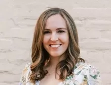 A woman with short hair is smiling while wearing a floral top in front of a white wall.
