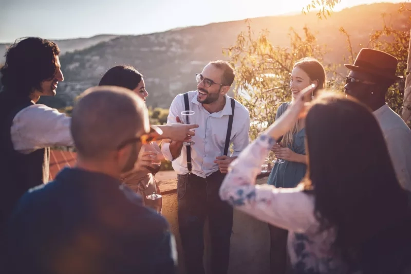 Group of people drinking wine at winery event with mountain view and warm sunset ambiance.