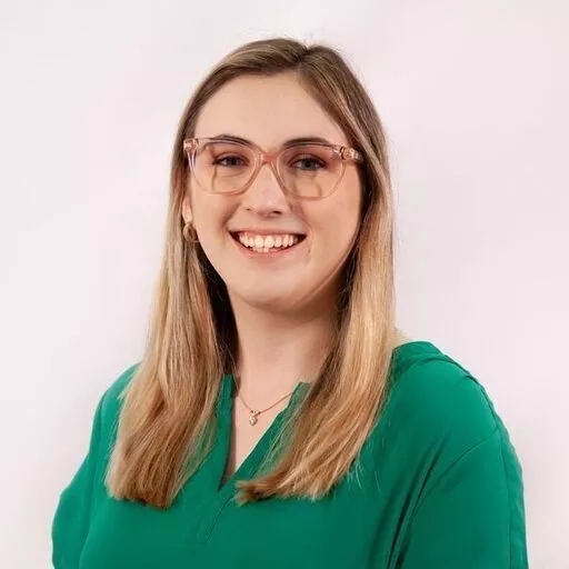 A smiling woman with glasses and a green shirt stands against a plain white wall.
