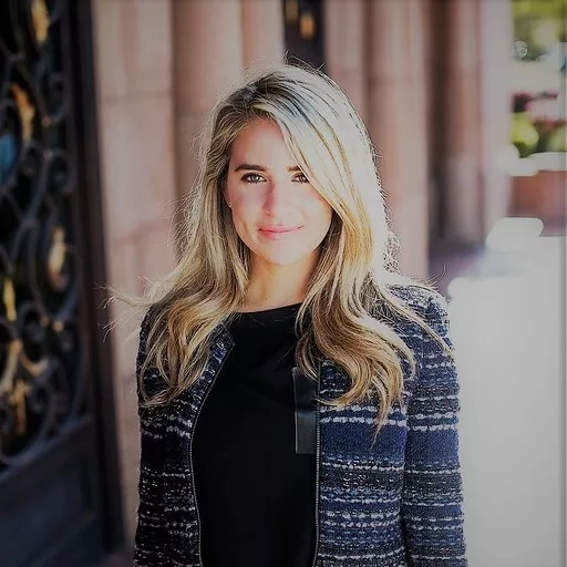 A smiling woman with blonde hair is standing in front of a building with an iron gate.
