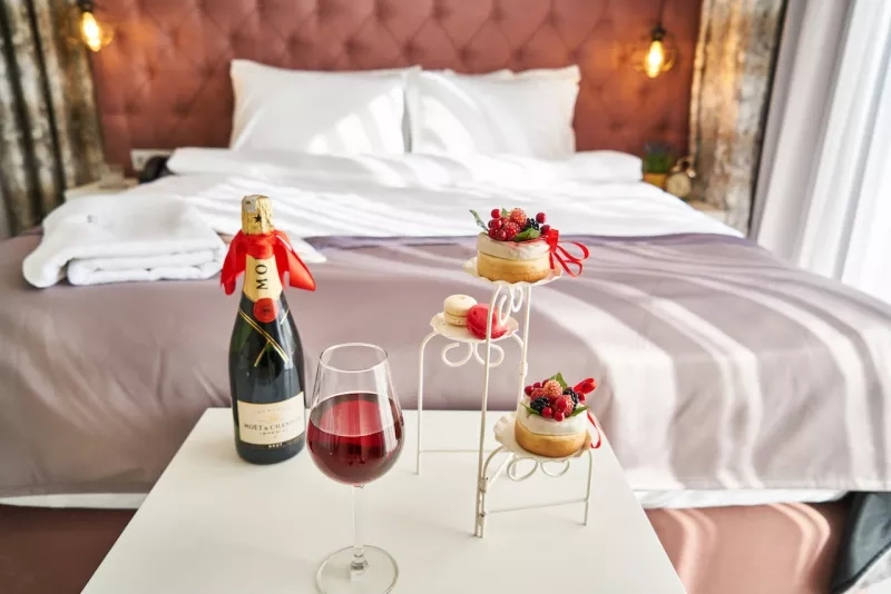 A champagne bottle, wine glass, and desserts are on a table in front of a bed.