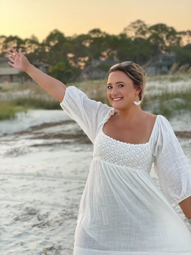 A woman in a white dress standing on a beach with her arm raised and smiling.