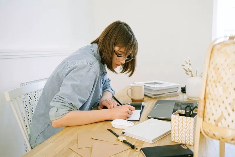 A woman is writing on a notepad at a table with a laptop and other stationery items.