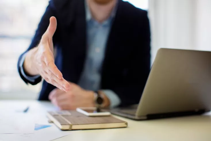 Businessman with laptop and phone on desk stretching hand forward in greeting or to shake hands.