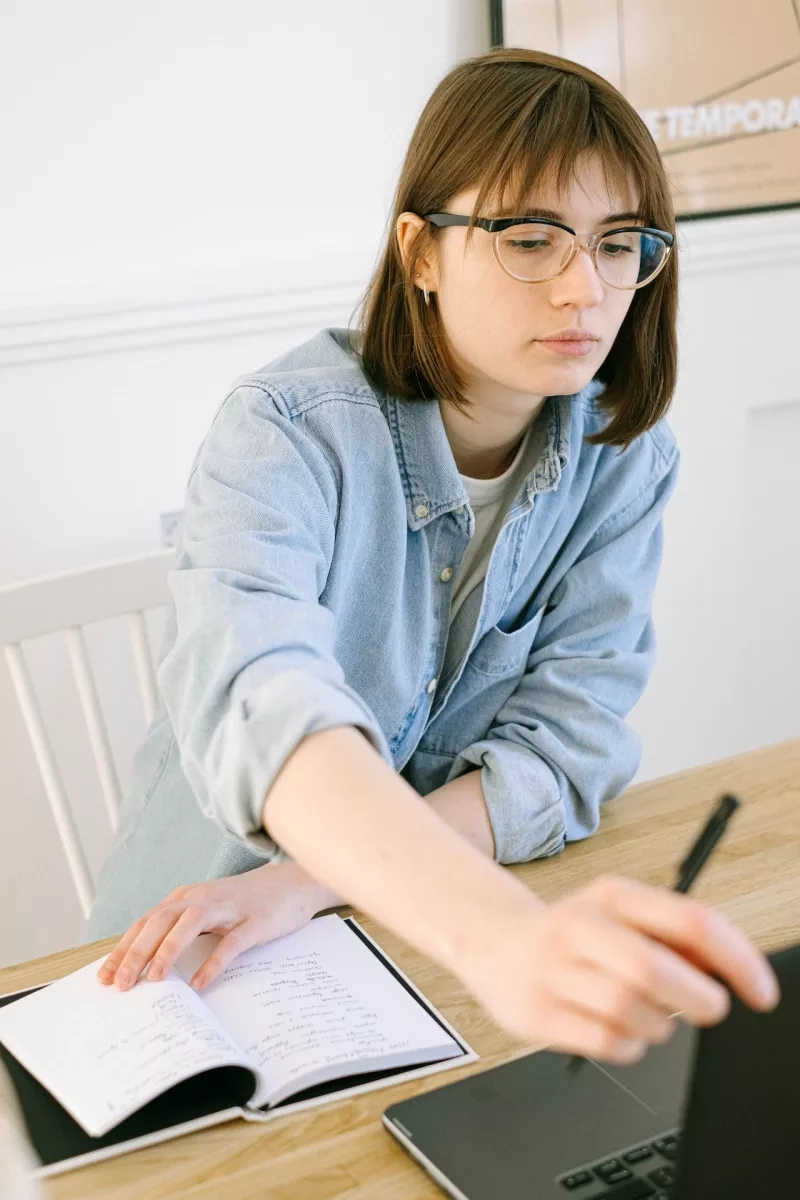 A woman with glasses on a laptop writing on a piece of paper with a pen.