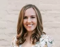JK Bio Pic of a lady with a smiling face standing against the white brick wall