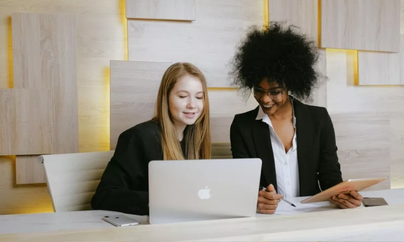 Two women sit at a desk with a laptop and tablet, working in a modern office setting.
