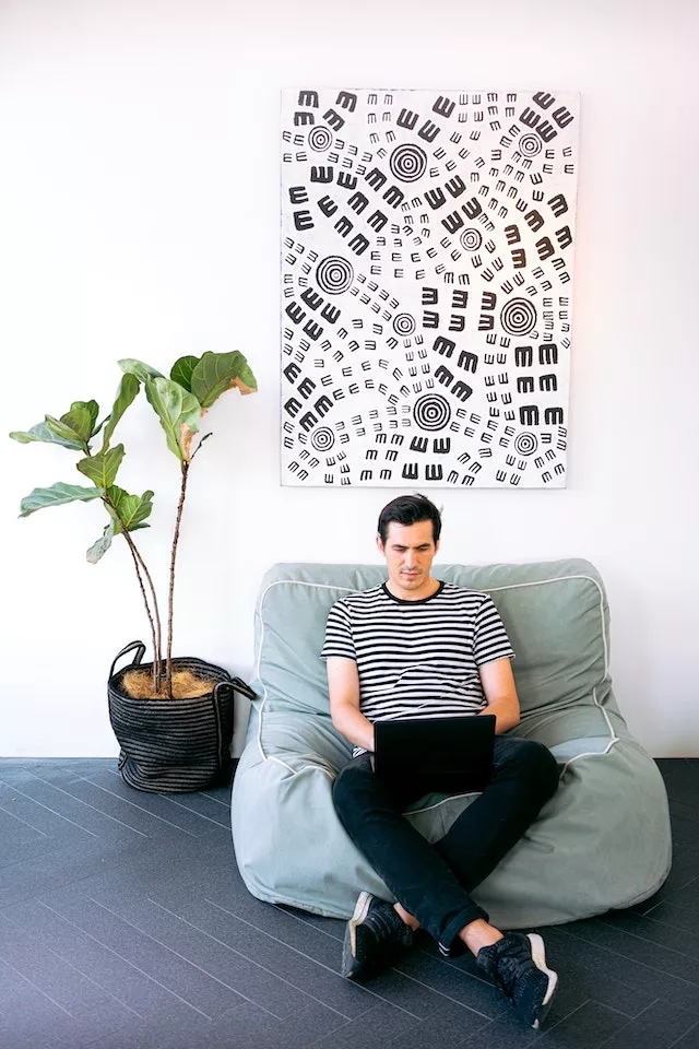 Man sitting on a beanbag chair working on his laptop in a modern, minimalist interior with a potted plant.