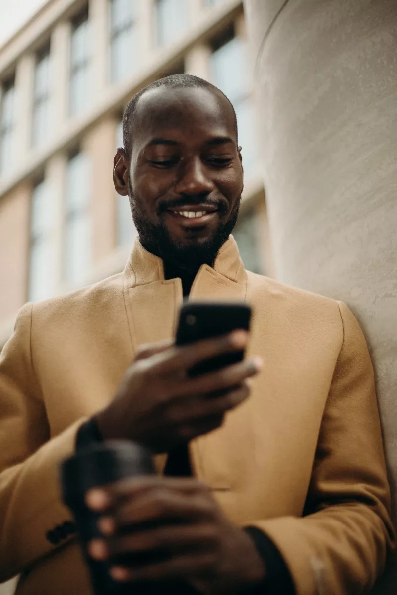 A man is smiling while holding a cell phone and standing in front of a building.