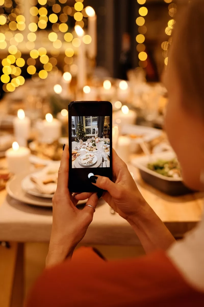 Person taking picture of table with candles and food in background of blurry yellow lights.