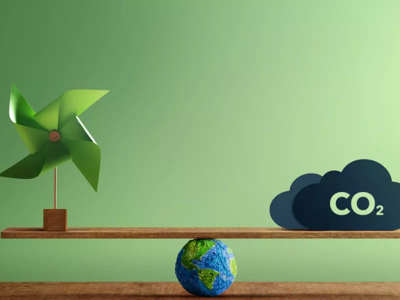 Pinwheel and earth model balancing on a seesaw with a CO2 cloud on the right side.