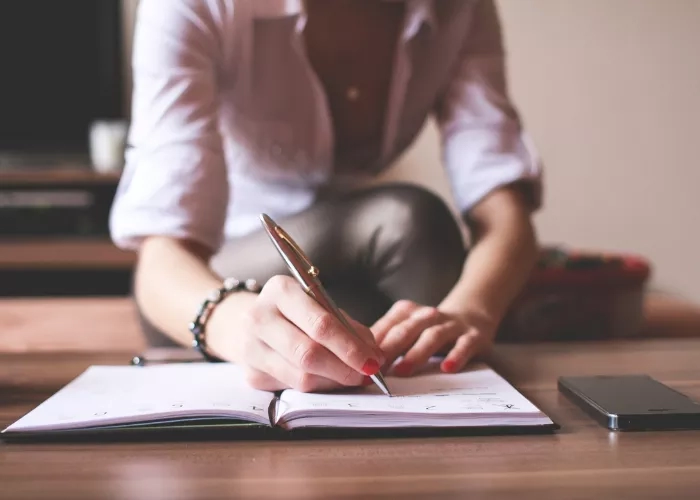 Woman writing in notebook with pen and cell phone on table, blurred background of TV and couch.