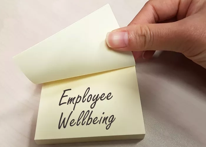 A hand holding a sticky note with the words Employee Wellbeing written on it.