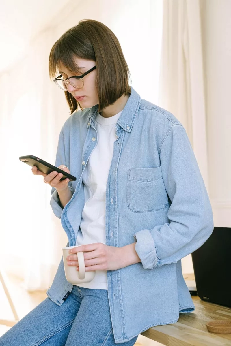 A woman with glasses in a denim jacket and jeans is sitting down with a smartphone.