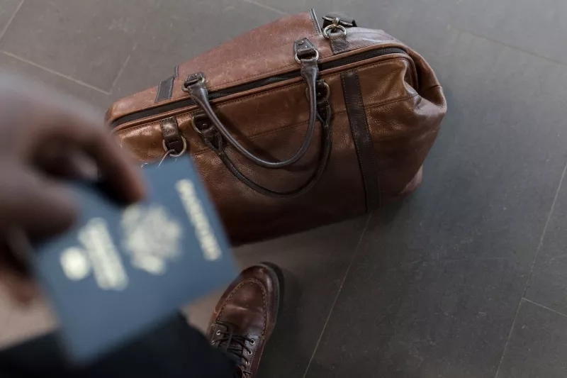 Person holding passport next to a leather bag with feet visible, tiled floor with dark grout.