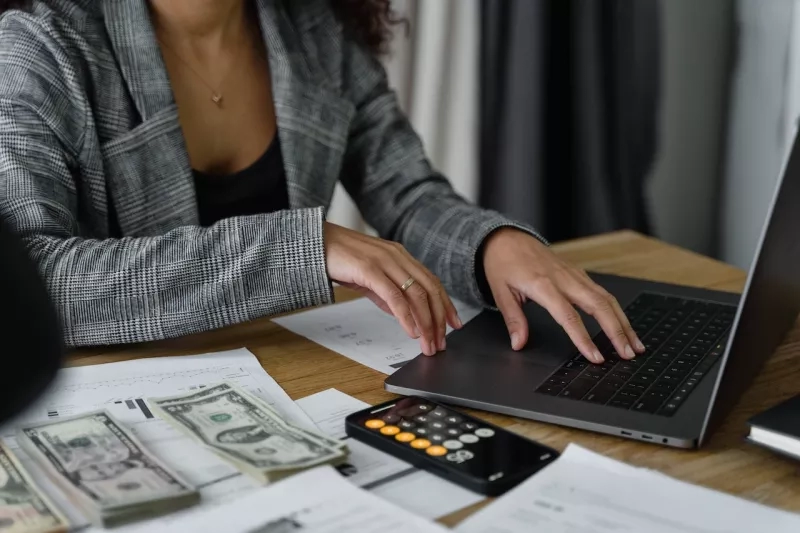 A woman is sitting at a desk typing on a laptop with a calculator and money on the desk.