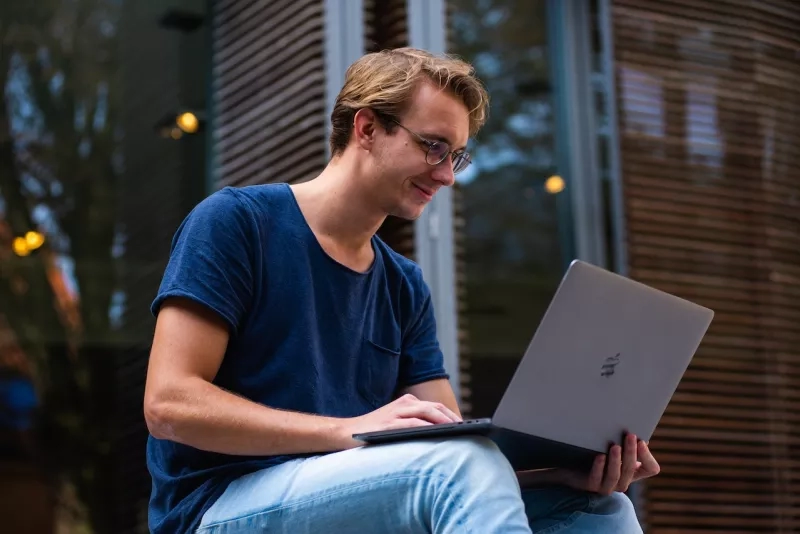 A man sitting outside with an Apple laptop on his lap, wearing a blue t-shirt and jeans.