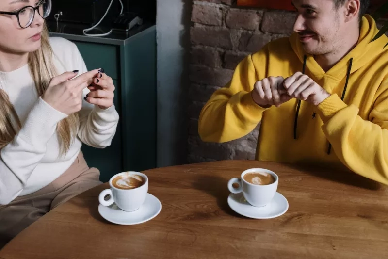 A man and a woman are sitting at a table with coffee cups, communicating using sign language.