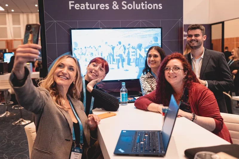 A group of people sitting at a table in front of a screen with the text "Features & Solutions."