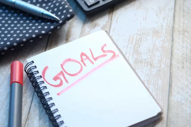 Notebook with the word goals written on it in pink marker on a wooden table with other office supplies.