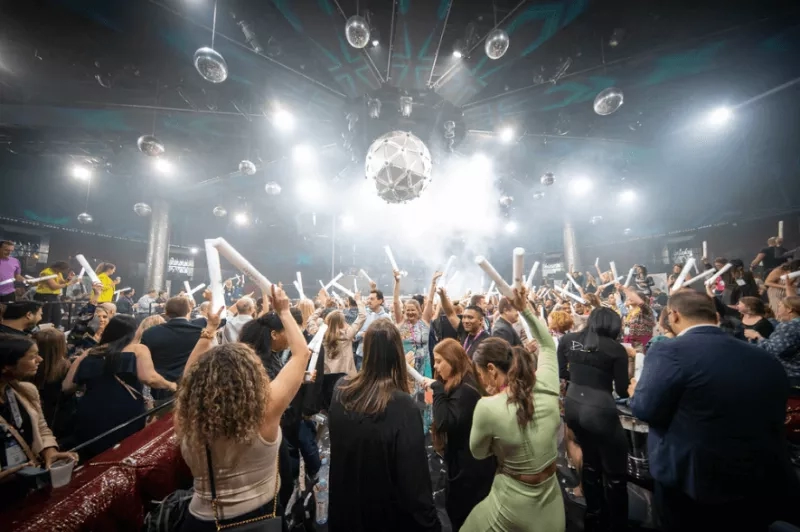 Crowded club with people raising white tubes in the air and a disco ball above them.