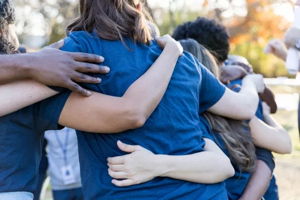 Group of people hug in a circle with arms around each other's shoulders in a park.