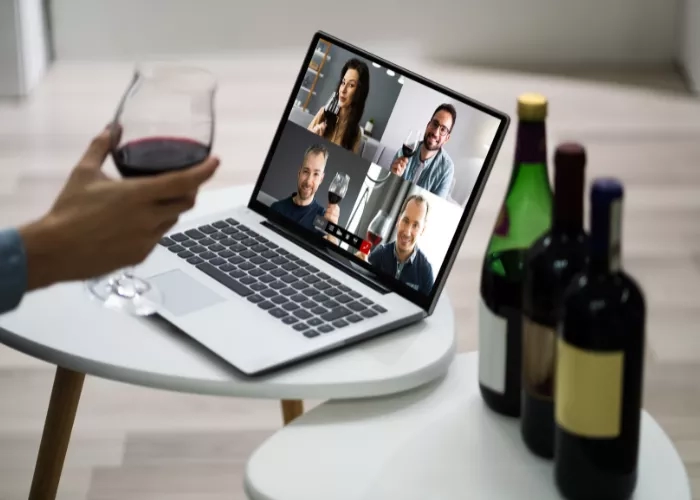 A laptop with four people on screen holding wine glasses, with wine bottles next to it.
