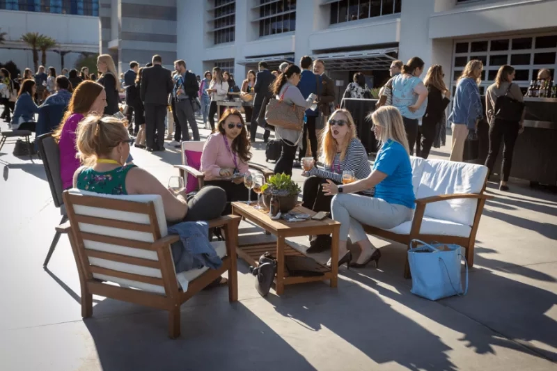 Four women sitting outdoors on chairs at an event with wine glasses on a table in front of them.