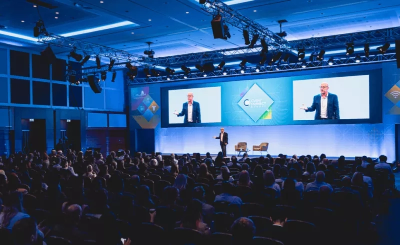 Two men giving speeches at a conference, with an audience watching them and screens behind them.