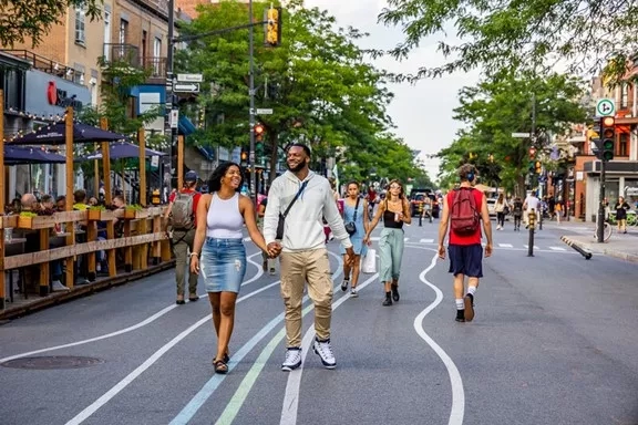 Diverse group of people walking down a busy city street with storefronts, trees, and traffic signals.