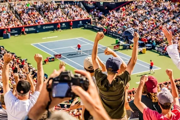Crowd cheering in stands at Rogers Cup, with two players on court, one holding camera, and one raising cap.