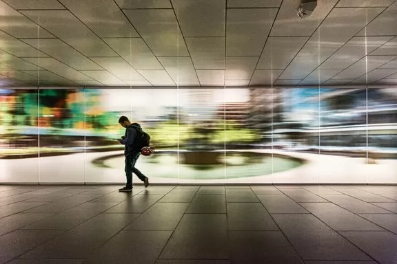 A man walking in an indoor hallway with a large blurred painting of a park in the background.