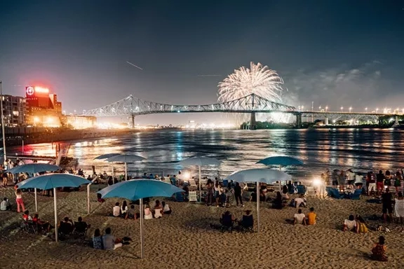 A crowd of people at the beach with fireworks and umbrellas during the summer season.
