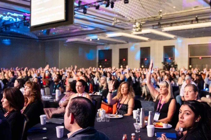 Large group of people sitting in a conference hall with a projector and cups on tables.