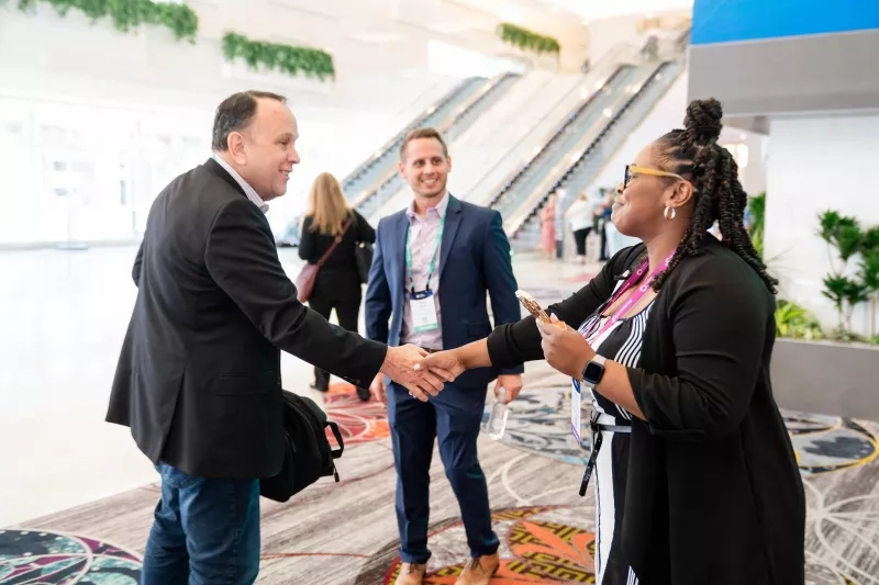 Two people shaking hands at a conference while a man looks on from behind them.