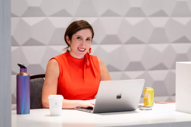 Woman in an orange blouse working on a laptop at an event with drinks on the table.