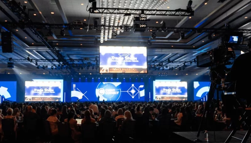 A large conference room with Trade Show banners and a crowd of people in the seats.