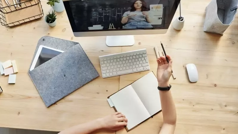 A person sitting at a desk with a notebook and a computer screen with a woman on it.