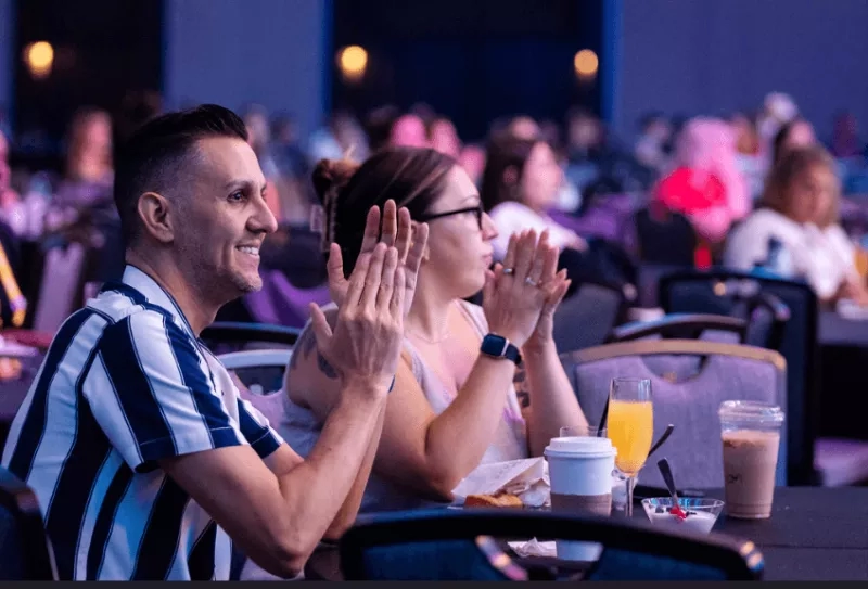 Two people clapping hands while sitting on chairs with food and drinks on the table in front of them.