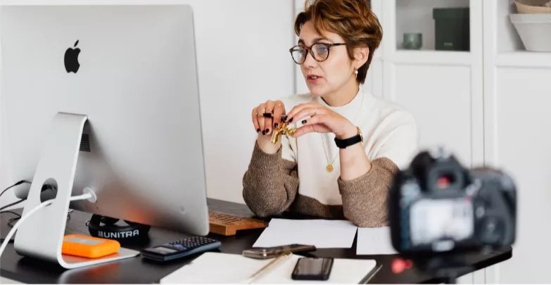 A woman with curly hair is sitting on a desk in front of an Apple computer.