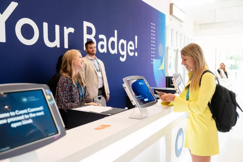 Two women are at a counter with a sign that says "Your Badge!" at a Cvent event.