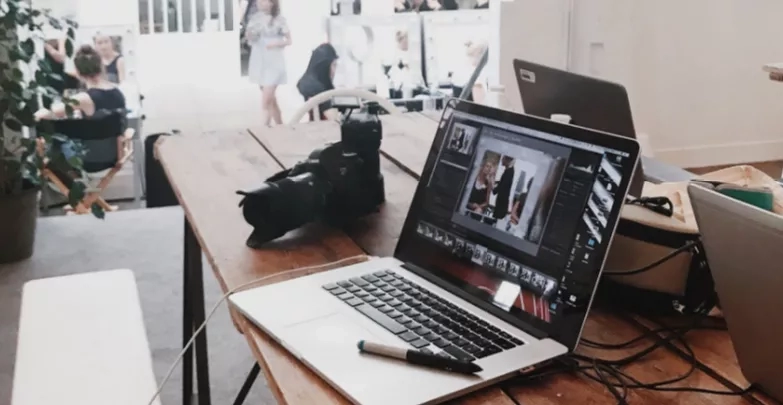 Laptop on wooden table with pen and camera, people sitting and walking in background.