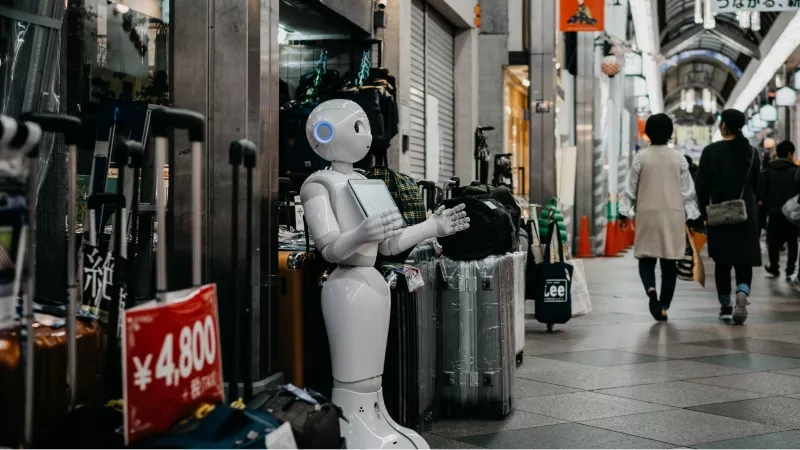 A white robot stands beside a store in a busy street with people walking behind.
