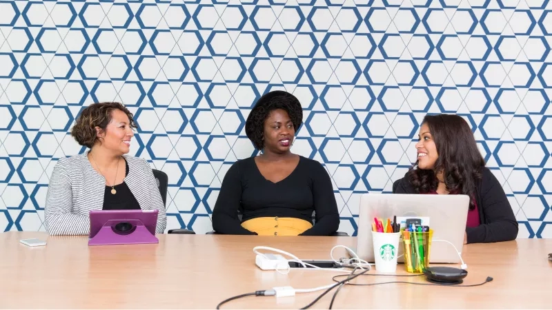Three women sit at a conference table with a laptop and tablet, smiling at the camera.