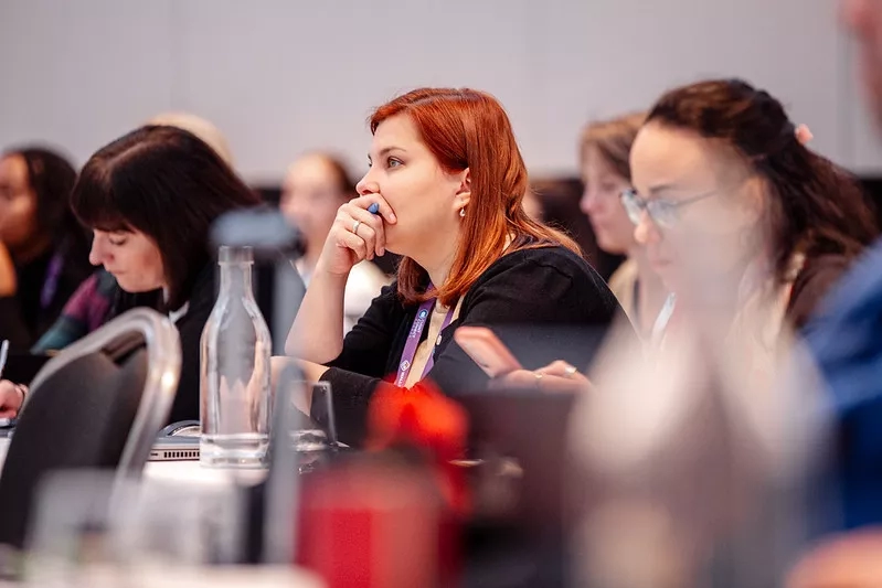 A group of women in a meeting with one woman using a pen to tap on her cheek.